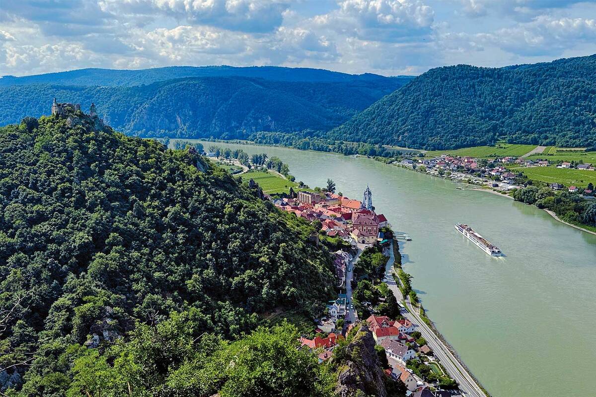 Die Bilderbuchkulisse der Wachau bei Dürnstein mit dem markanten blauen Kirchturm und der Burgruine auf dem Felsen, während ein Kreuzfahrtschiff gemächlich die Donau hinabgleitet.